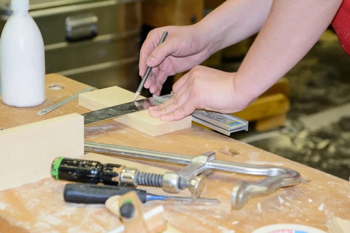 Hands squaring a small wooden workpiece on a bench beside a clamp, a chisel and a marking gauge.