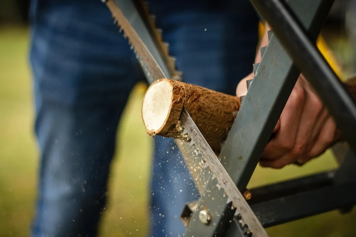 A person cutting a log with a bow saw supported on a folding sawhorse outdoors.