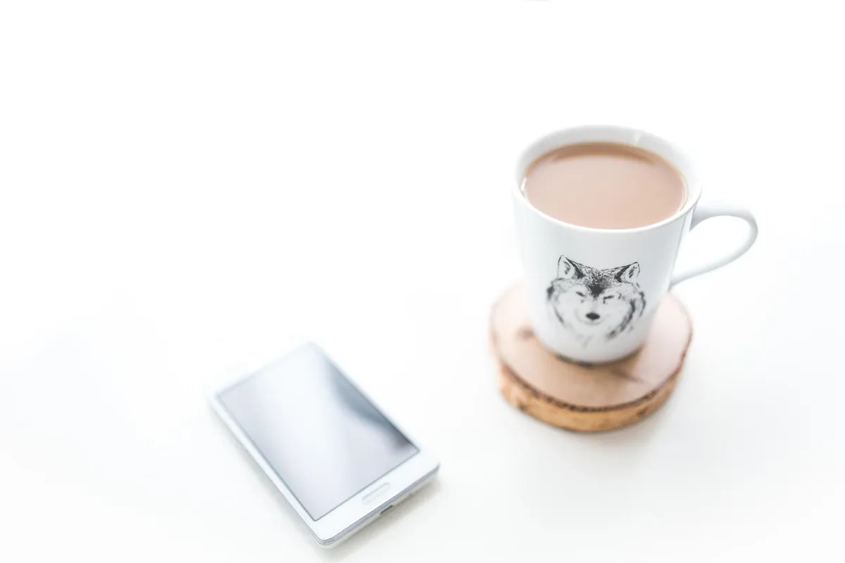 A mug of coffee resting on a round wooden coaster on a white desk beside a phone.