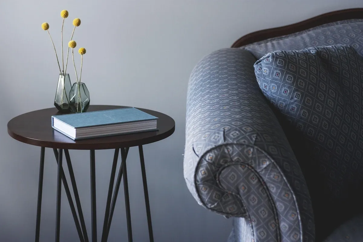 A small round side table holding a book and a vase, standing beside an upholstered armchair.