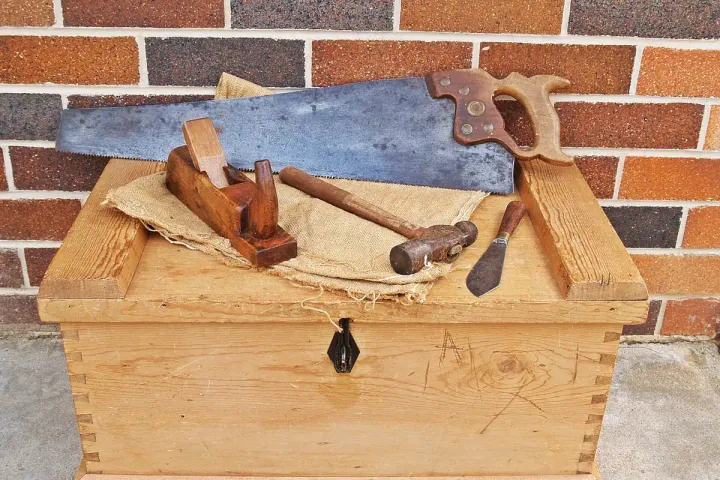 A handmade wooden toolbox topped with a hand saw, wooden block plane, hammer and chisel against a brick wall.
