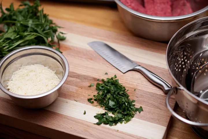 A wooden chopping board on a kitchen counter with a knife and chopped herbs on its surface.