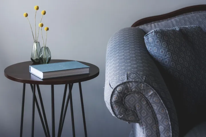 A small round side table holding a book and a vase, standing beside an upholstered armchair.